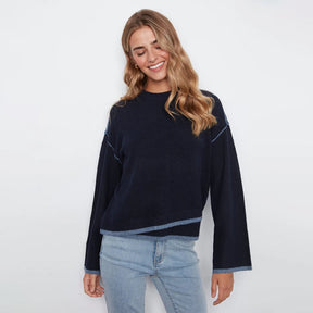 A woman wearing a dark navy blue knit sweater with light blue contrast stitching and light-wash blue jeans, standing against a plain white studio background.
