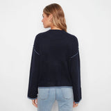 A woman viewed from the back wearing a dark navy knit sweater with light blue contrast stitching on the shoulders and light-wash blue jeans, against a plain white studio background.