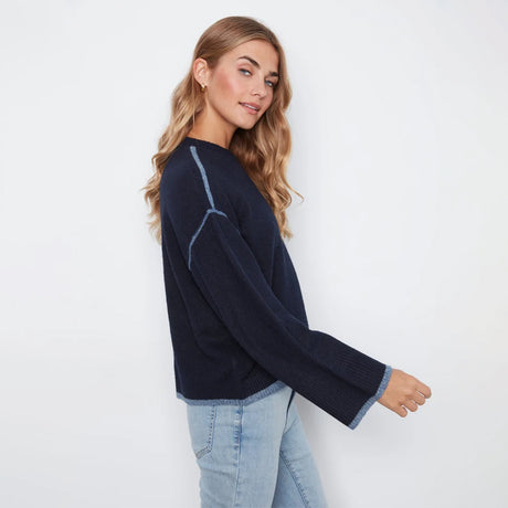 A woman wearing a dark navy blue knit sweater with light blue contrast stitching and light-wash blue jeans, photographed against a plain white studio background.
