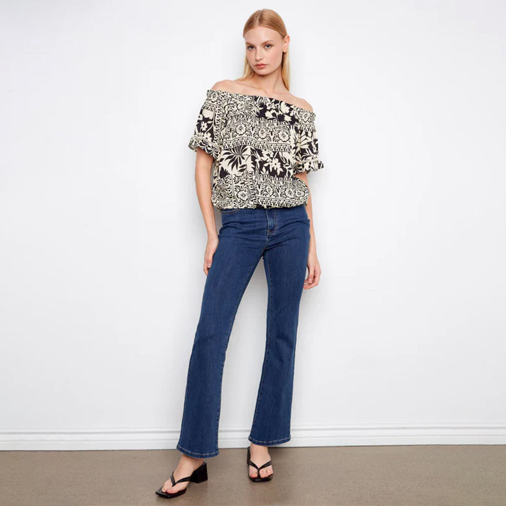 Woman wearing a floral off-shoulder top and blue jeans against a white wall.