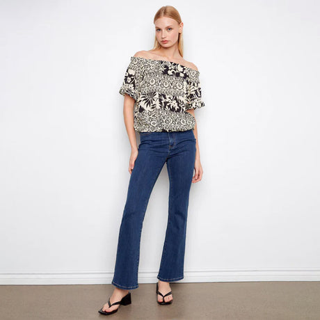 Woman wearing a floral off-shoulder top and blue jeans against a white wall.