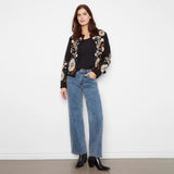 A woman stands against a plain white wall wearing a black floral embroidered cardigan over a black top, blue wide-leg jeans, and black ankle boots.
