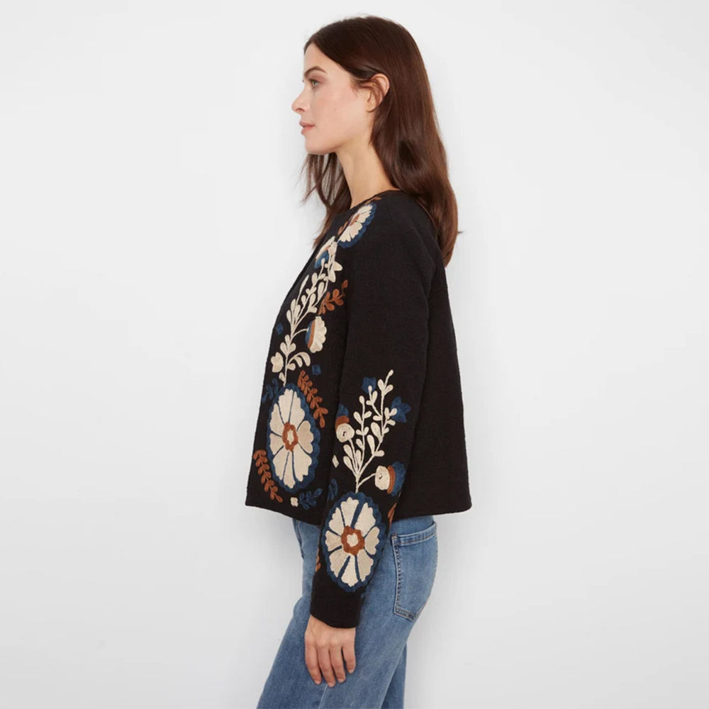 A woman in profile wearing a black knit sweater with large floral embroidery in cream, blue, and brown, paired with blue jeans, against a plain white background.