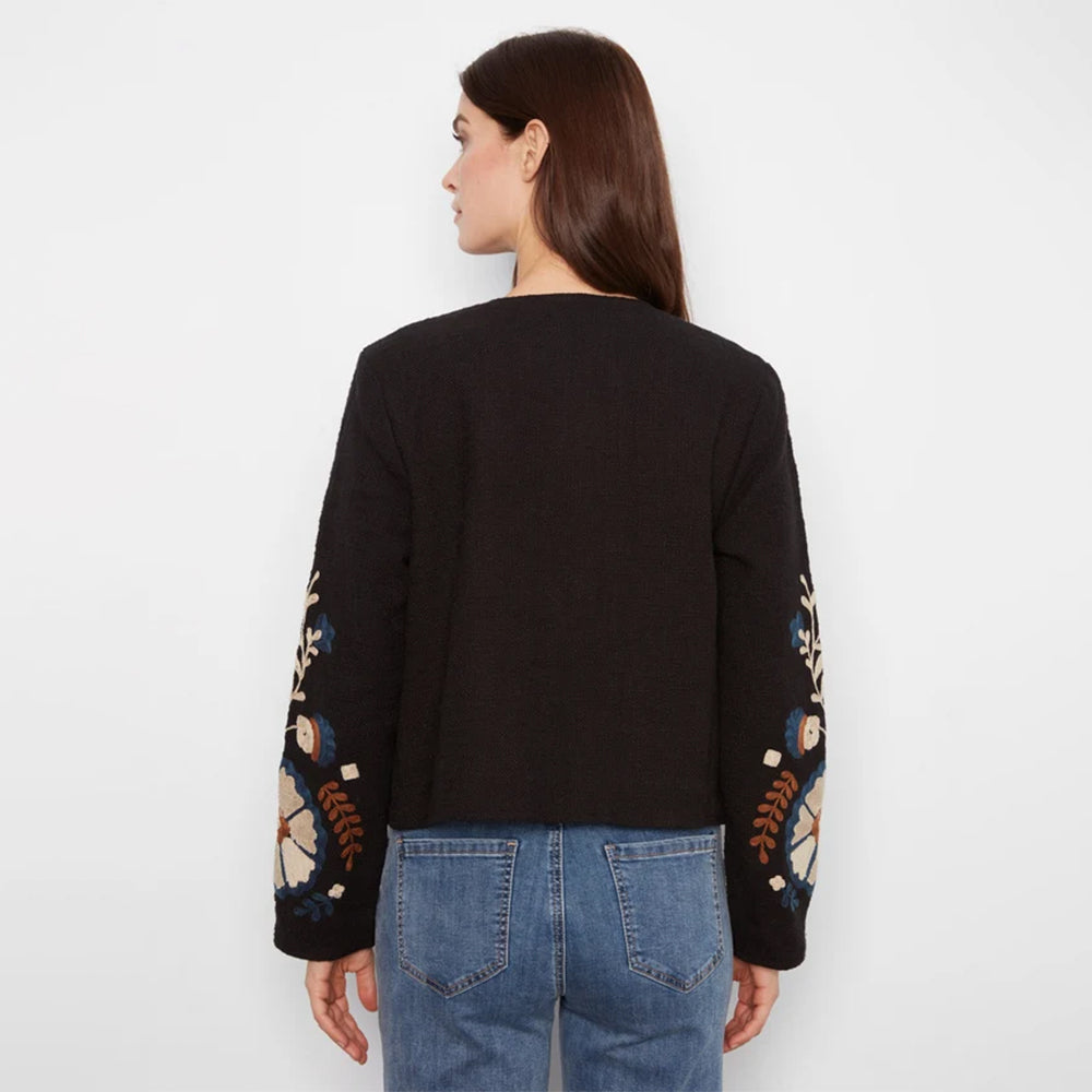 A woman viewed from the back wearing a black cropped sweater with colorful floral embroidery on the sleeves and blue jeans, against a plain white background.