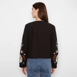 A woman viewed from the back wearing a black cropped sweater with colorful floral embroidery on the sleeves and blue jeans, against a plain white background.