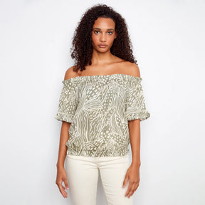 Woman wearing a patterned off-shoulder top and white pants on a plain background