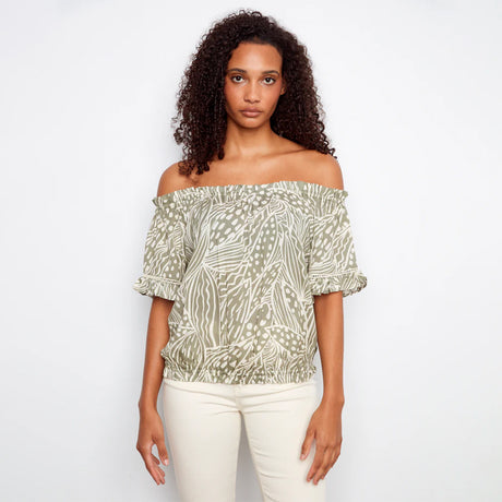 Woman wearing a patterned off-shoulder top and white pants on a plain background