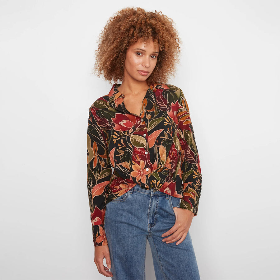A woman with curly light brown hair wearing a long-sleeve button-up blouse with a vibrant red, orange, and green floral pattern on a black background, paired with blue jeans, photographed against a light gray studio background.