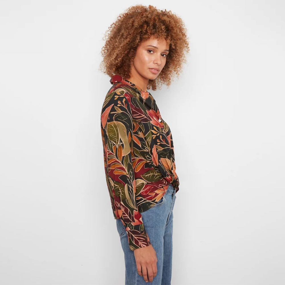 A woman with curly light brown hair is shown from the side, wearing a long-sleeve button-up blouse with a dense tropical leaf pattern in red, orange, green, and black, paired with blue jeans against a plain white background.