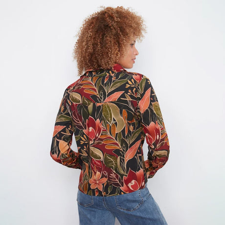 A woman with curly light brown hair is shown from the back wearing a long-sleeve button-up shirt with a dense tropical floral print in red, orange, green, and black, paired with blue jeans against a plain white background.