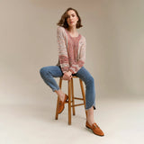 A woman sits on a wooden stool wearing a chunky knit long-sleeve sweater in a pink, red, and beige pattern, blue jeans, and tan loafers against a neutral beige studio background.