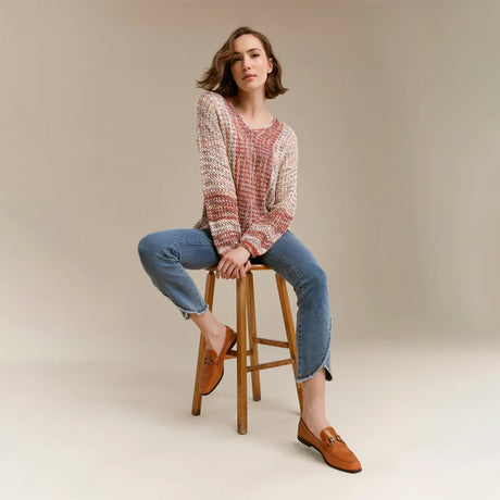 A woman sits on a wooden stool wearing a chunky knit long-sleeve sweater in a pink, red, and beige pattern, blue jeans, and tan loafers against a neutral beige studio background.