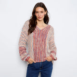 A woman modeling a chunky knit sweater with a multicolor pattern of red, blue, beige, and white yarn, paired with blue jeans, against a plain white background.