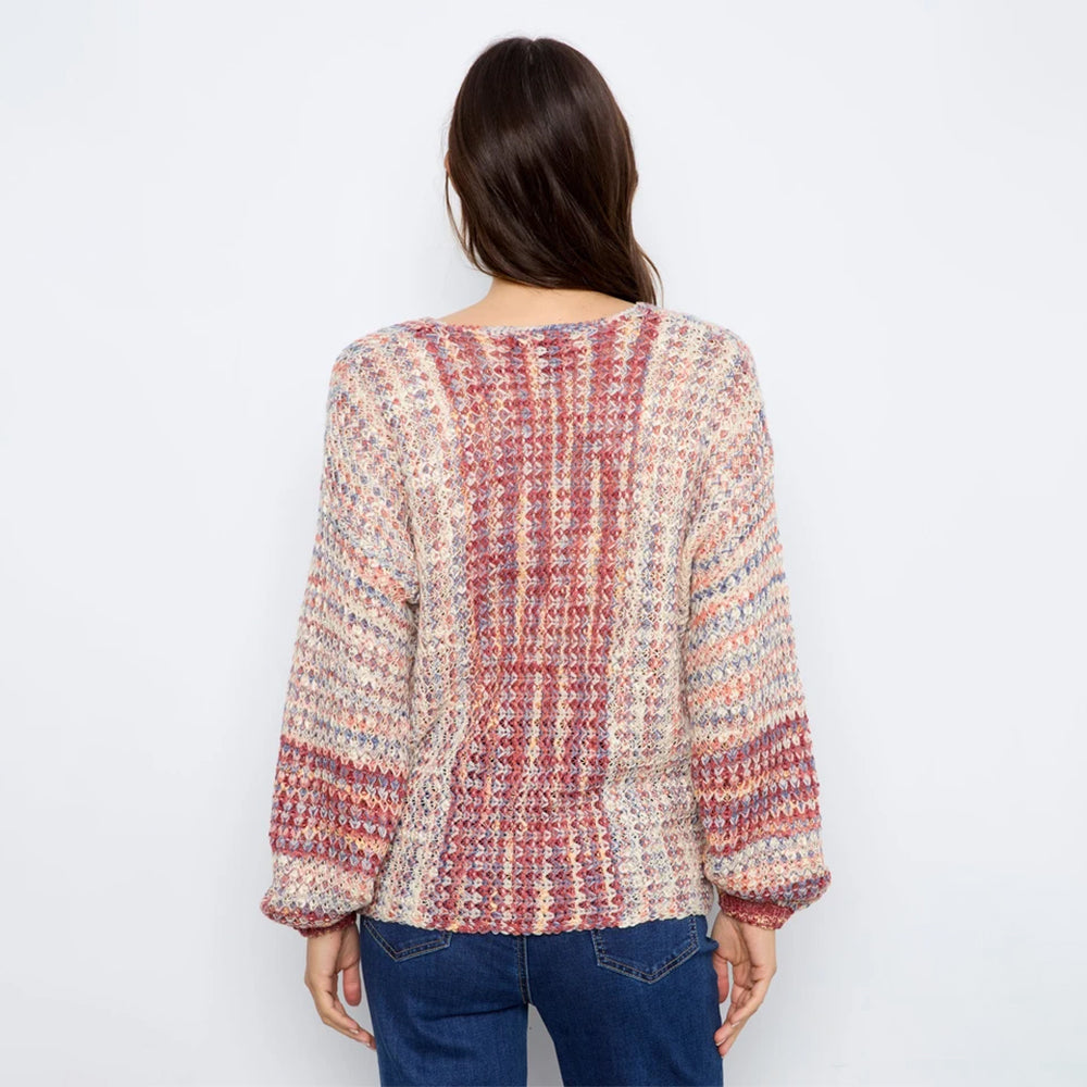 A woman viewed from behind wearing a chunky, multicolored knit sweater with blue jeans, set against a plain white studio background.