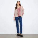 A woman stands against a plain white wall wearing a loose, multicolor knit sweater with blue jeans and dark brown leather loafers.