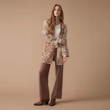 A woman models a mid-length open-front cardigan with an intricate beige and brown damask pattern, styled over a white top, brown wide-leg pants, and brown ankle boots against a solid beige studio backdrop.