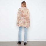 A woman viewed from behind wearing a beige cardigan with a brown floral pattern, paired with light blue jeans and black heels, standing against a white wall.