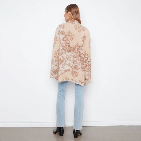 A woman viewed from behind wearing a beige cardigan with a brown floral pattern, paired with light blue jeans and black heels, standing against a white wall.