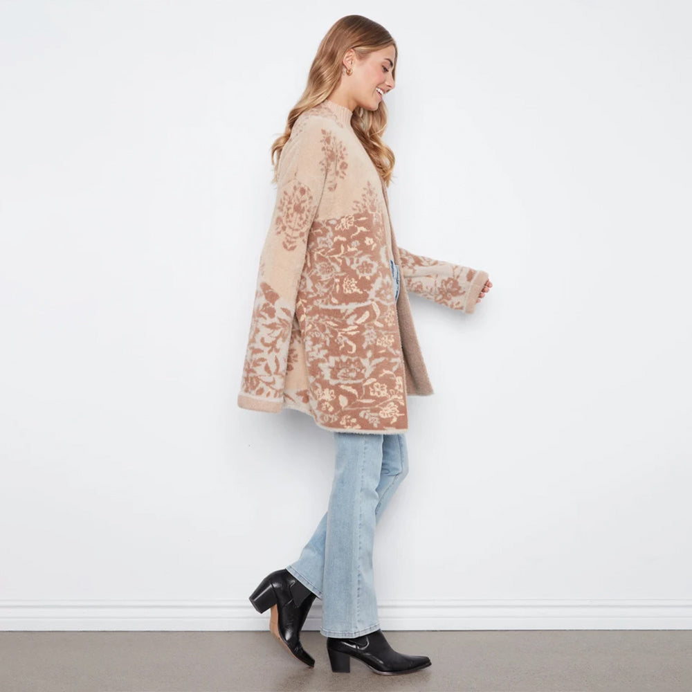 A woman wearing a beige and brown patterned cardigan, light blue jeans, and black ankle boots, standing against a white wall.