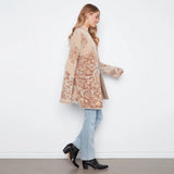 A woman wearing a beige and brown patterned cardigan, light blue jeans, and black ankle boots, standing against a white wall.