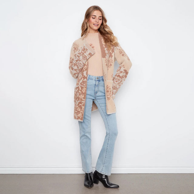 A woman modeling a beige open-front cardigan with brown floral lace-like patterns, worn over a ribbed beige top, light-wash blue jeans, and black ankle boots, standing against a plain white wall.
