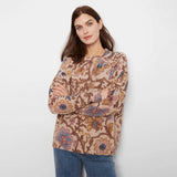 A woman modeling a long-sleeve crewneck sweater with a detailed floral and botanical pattern in shades of brown, beige, and blue, paired with blue jeans against a plain light gray background.
