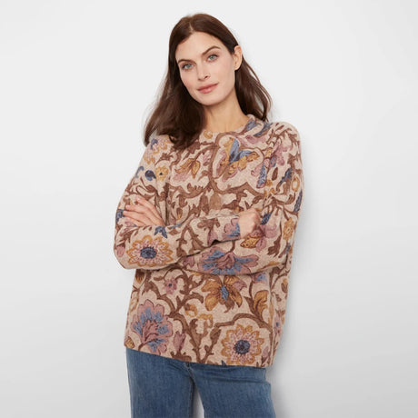 A woman modeling a long-sleeve crewneck sweater with a detailed floral and botanical pattern in shades of brown, beige, and blue, paired with blue jeans against a plain light gray background.