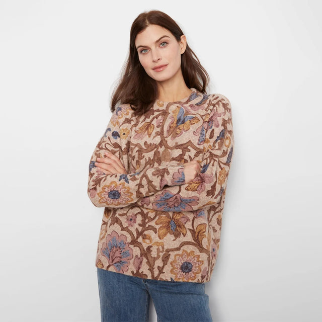 A woman modeling a long-sleeve crewneck sweater with a detailed floral and botanical pattern in shades of brown, beige, and blue, paired with blue jeans against a plain light gray background.