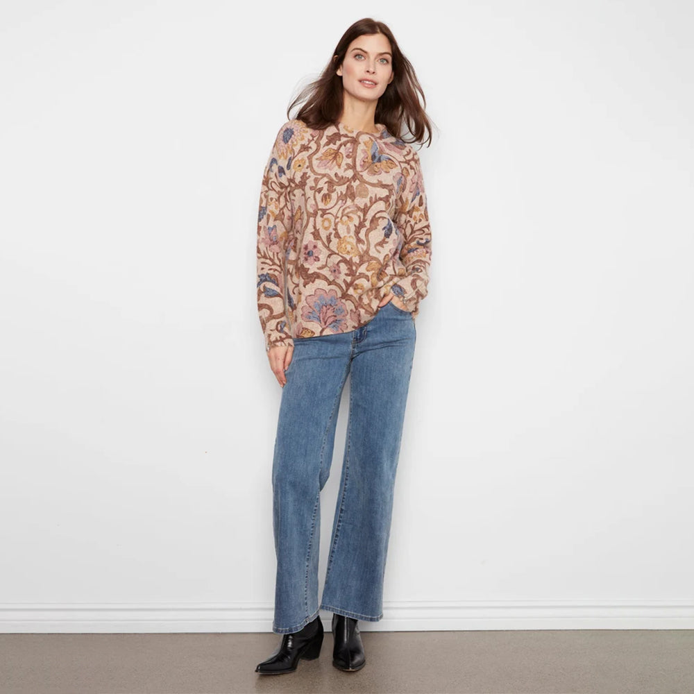 A woman modeling a long-sleeve crewneck sweater with a detailed floral and botanical pattern in shades of brown, beige, and blue, paired with blue jeans against a plain light gray background.