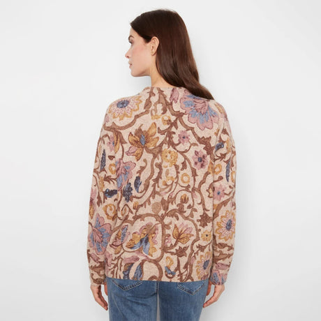 A woman shown from the back wearing a beige long-sleeve knit sweater with a dense floral and botanical pattern in brown, pink, blue, and yellow, paired with blue jeans against a plain white background.