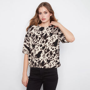 A woman modeling a short-sleeve crewneck top with a black and beige cow-print pattern, paired with black jeans, against a plain white studio background.