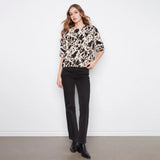 A woman stands against a white wall wearing a beige and black abstract-patterned short-sleeve top, black pants, and black ankle boots.