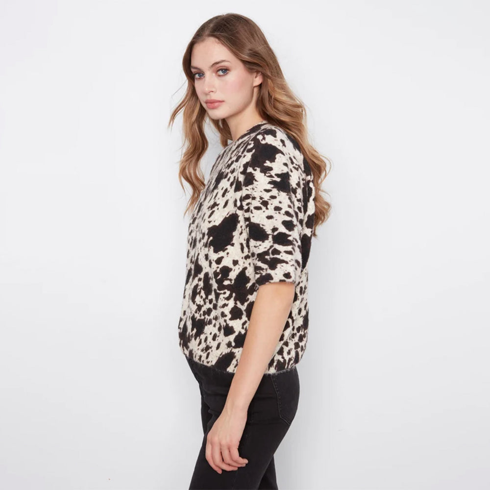 A woman wearing a short-sleeve knit top with a black and beige cow-print pattern, paired with black pants, photographed against a plain white studio background.