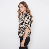 A woman wearing a short-sleeve knit top with a black and beige cow-print pattern, paired with black pants, photographed against a plain white studio background.