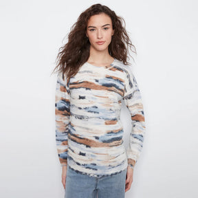 A woman with long, wavy brown hair wearing a long-sleeve knit sweater with an abstract watercolor pattern in beige, brown, blue, and white, paired with light-wash blue jeans, against a plain white studio background.