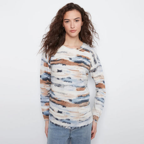 A woman with long, wavy brown hair wearing a long-sleeve knit sweater with an abstract watercolor pattern in beige, brown, blue, and white, paired with light-wash blue jeans, against a plain white studio background.