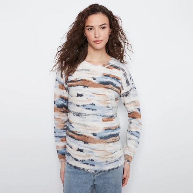 A woman with long, wavy brown hair wearing a long-sleeve knit sweater with an abstract watercolor pattern in beige, brown, blue, and white, paired with light-wash blue jeans, against a plain white studio background.