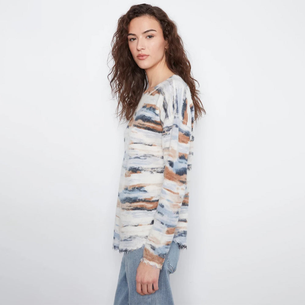 A woman modeling a long-sleeve knit sweater with a distressed, watercolor-like abstract pattern in cream, blue, and brown, paired with light blue jeans against a plain white studio background.
