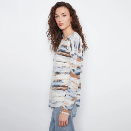 A woman modeling a long-sleeve knit sweater with a distressed, watercolor-like abstract pattern in cream, blue, and brown, paired with light blue jeans against a plain white studio background.