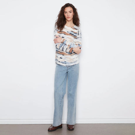 A woman stands against a white wall wearing a long-sleeve knit sweater with an abstract brushstroke pattern in cream, blue, brown, and gray, paired with light-wash wide-leg jeans and dark brown loafers.