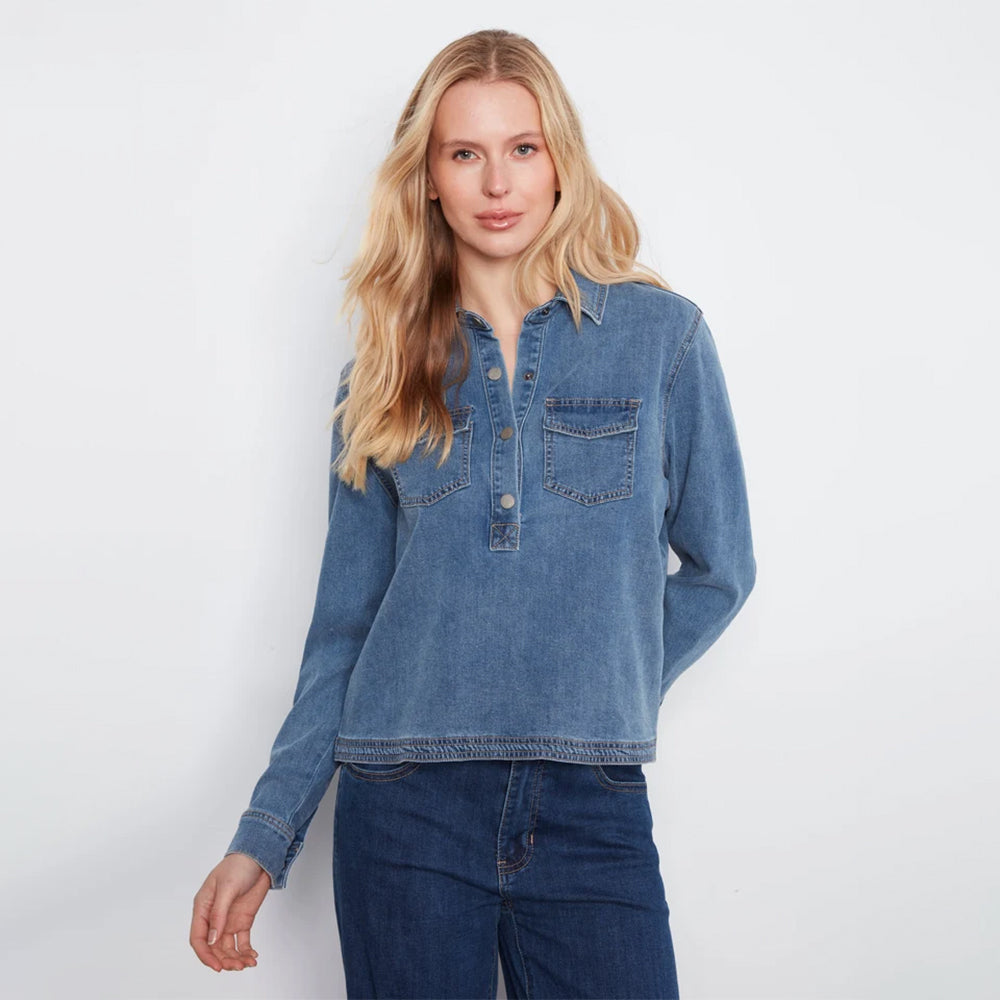 A woman stands against a plain white wall wearing a light-wash blue denim long-sleeve button-up shirt and dark blue wide-leg jeans.