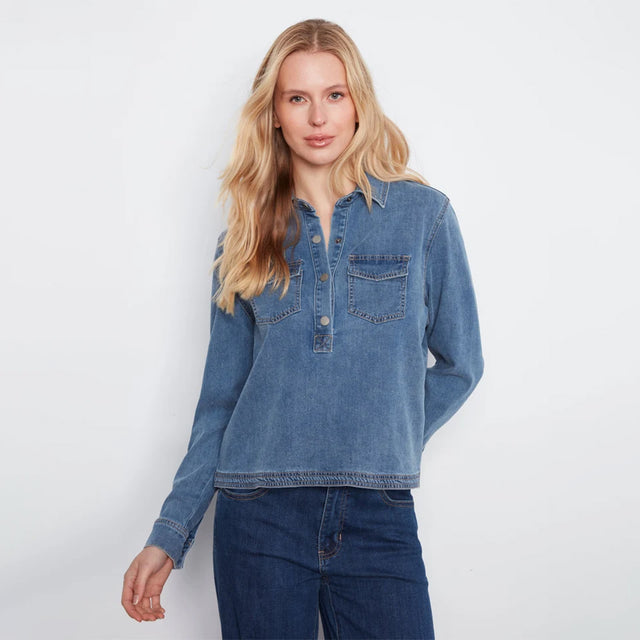 A woman stands against a plain white wall wearing a light-wash blue denim long-sleeve button-up shirt and dark blue wide-leg jeans.