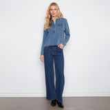 A woman stands against a plain white wall wearing a light-wash blue denim long-sleeve button-up shirt and dark blue wide-leg jeans, paired with black shoes.