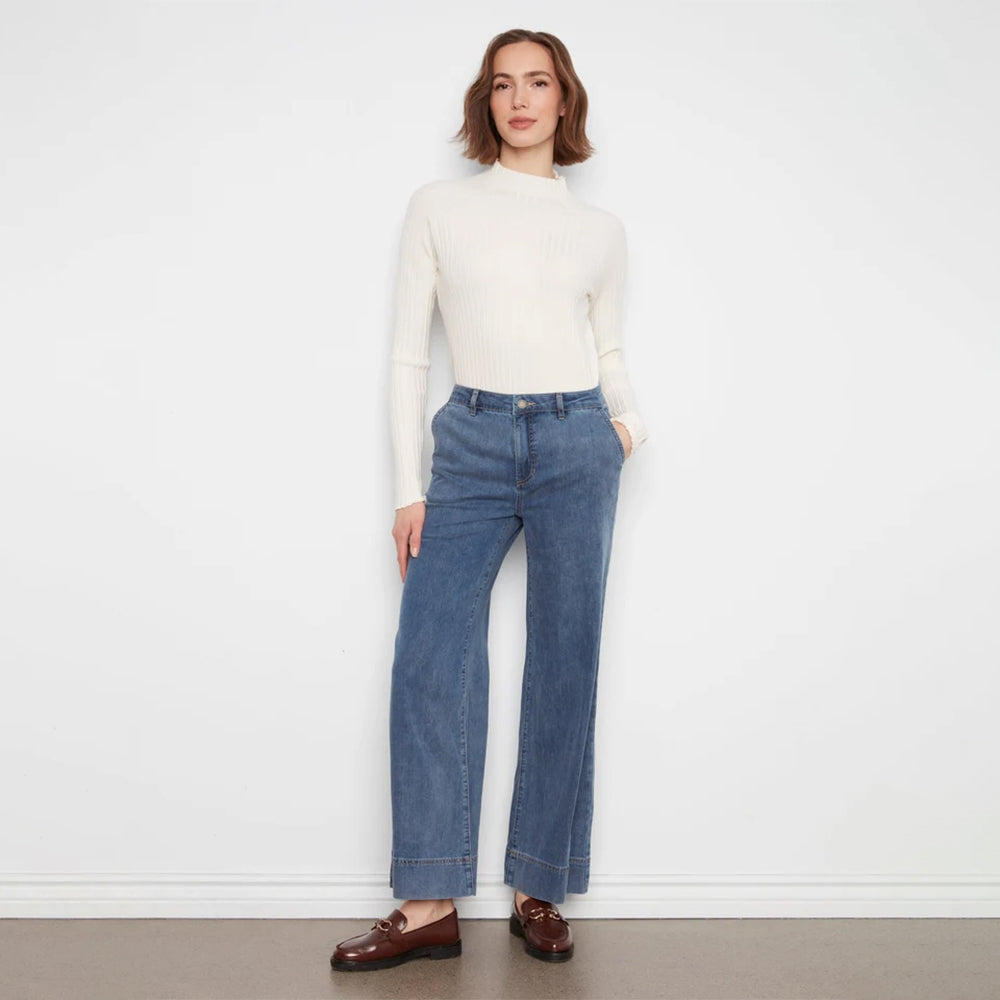 A woman stands against a plain white wall wearing a cream ribbed mock-neck long-sleeve top, blue wide-leg jeans, and brown leather loafers.