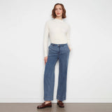 A woman stands against a plain white wall wearing a cream ribbed mock-neck long-sleeve top, blue wide-leg jeans, and brown leather loafers.