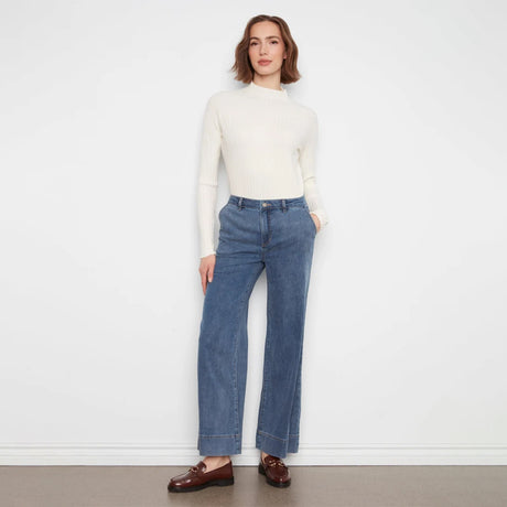A woman stands against a plain white wall wearing a cream ribbed mock-neck long-sleeve top, blue wide-leg jeans, and brown leather loafers.