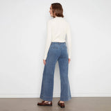 A woman is shown from the back wearing a cream mock-neck long-sleeve top, blue wide-leg cropped jeans, and brown leather loafers, standing against a white wall.