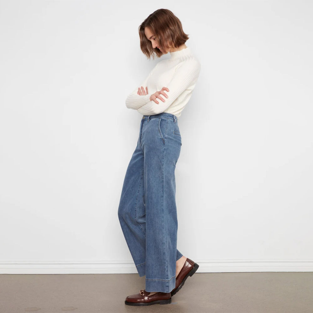 A woman stands in profile wearing a white mock-neck knit sweater, wide-leg blue jeans, and brown leather loafers against a plain white wall.