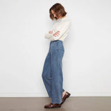 A woman stands in profile wearing a white mock-neck knit sweater, wide-leg blue jeans, and brown leather loafers against a plain white wall.