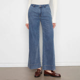 A model wearing a pair of wide-leg blue denim jeans with a rolled hem, styled with a cream ribbed long-sleeve top and brown leather loafers against a white wall and gray floor.
